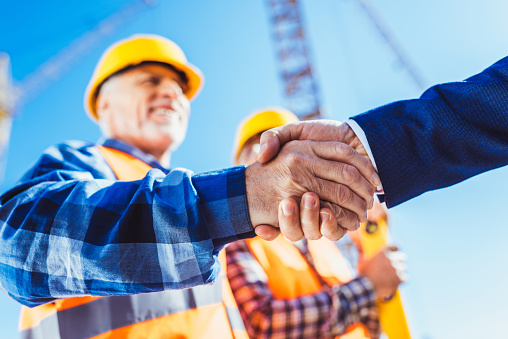 Construction worker in protective uniform shaking hands with businessman at construction site