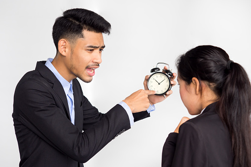 Asian businessman showing alarm clock and complaining at employee about working late on white background, punctuality concept