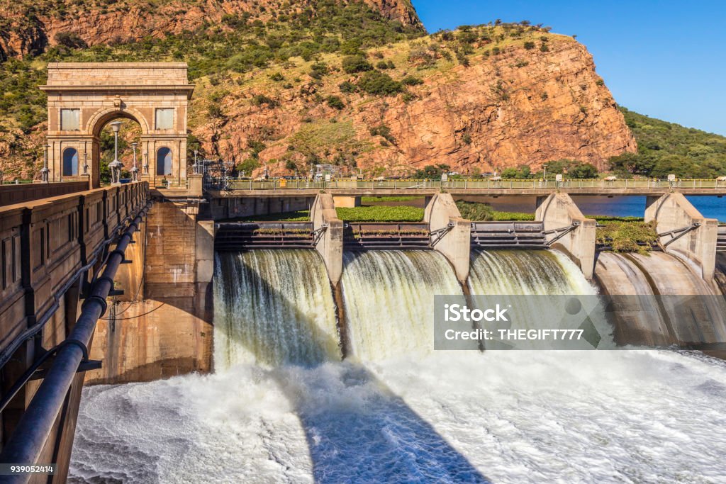 Hartbeespoort Dam and the wall panoramic Hartbeespoort Dam and the reservoir wall with the water gushing through the open gates of the reservoir after heavy rainfall. The iconic tower and single road allowing traffic to pass through with a tunnel through the cliff. This is a popular travel getaway for tourists from Johannesburg and Pretoria as is is situated within 45 minutes from the city centre. Hartbeespoort Dam Stock Photo