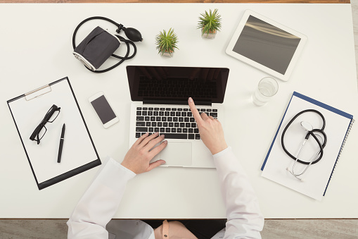 Doctor sitting at office desk with medical equipment and pointing on his laptop with blank screen, top view, copy space