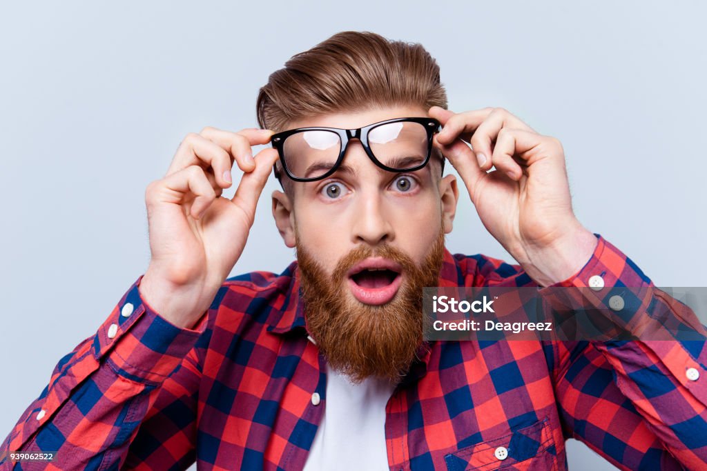 It's incredible! Close up portrait of young bearded man touching the spectacles and keeping his mouth open against gray background Men Stock Photo