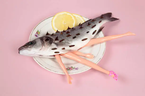 A bass fish with arms and legs of a doll inside on a flower plate. Cannibalism and anthropomorphism on a pink feminine background. quirky minimal color still life photography."n
