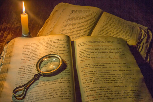 Opened an ancient prayer book and a magnifying glass on its open pages against the background of closed old books under the light of a candle.