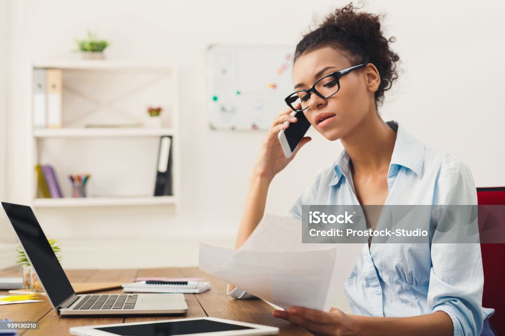 Serious woman consulting by phone at office Young serious african-american businesswoman talking by phone with papers, sitting at modern office workplace. Business consulting, copy space Using Phone Stock Photo