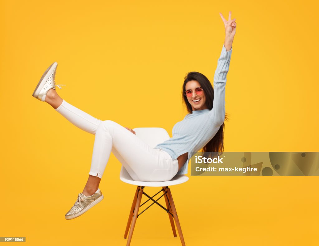 Excited hipster girl posing on chair Bright female in sunglasses sitting happily on chair in studio showing two fingers. Sitting Stock Photo