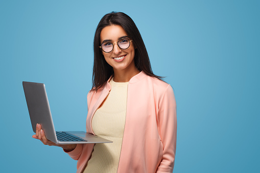 Young casual brunette in eyeglasses holding laptop and smiling at camera on blue.