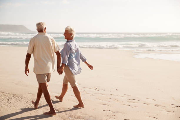 vue arrière du couple de personnes âgées marchant main dans la main le long de la plage - retirement beach women vacations photos et images de collection