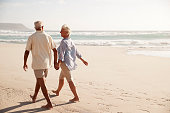 Rear View Of Senior Couple Walking Along Beach Hand In Hand
