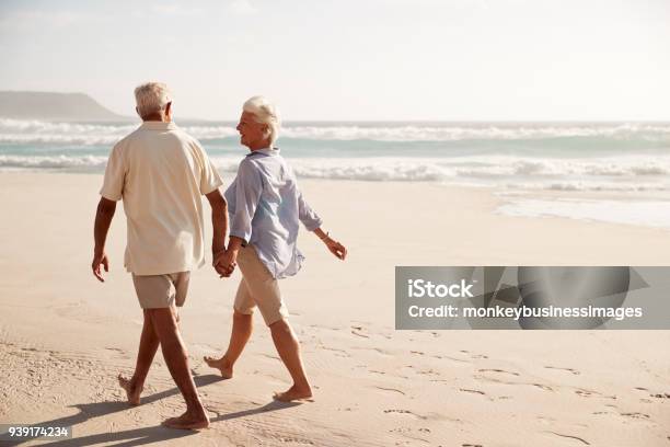 Rear View Of Senior Couple Walking Along Beach Hand In Hand Stock Photo - Download Image Now