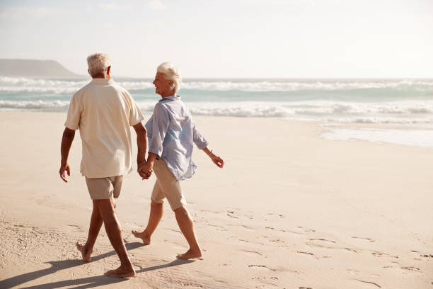 задний вид старшей пары, идущей вдоль пляжа рука об руку - senior adult senior couple couple summer стоковые ф�ото и изображения