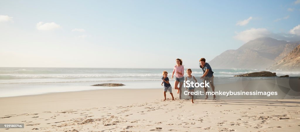 Genitori che corrono lungo la spiaggia con i bambini durante le vacanze estive - Foto stock royalty-free di Famiglia
