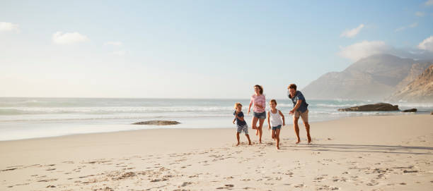 genitori che corrono lungo la spiaggia con i bambini durante le vacanze estive - chasing women men couple foto e immagini stock