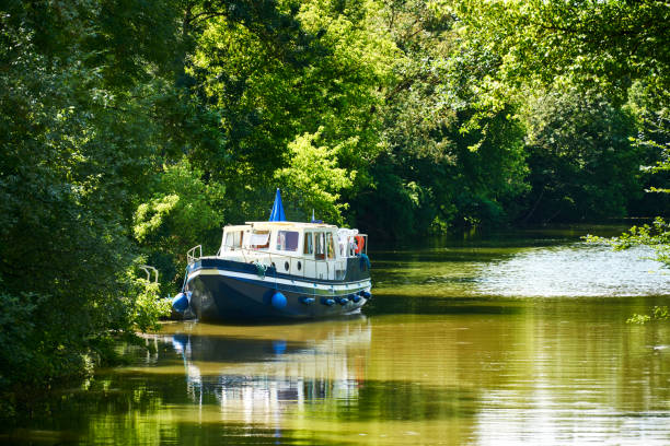 Houseboat anchors on the shore of a canal with trees in summer