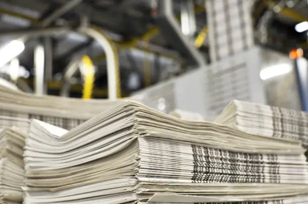 Photo of stack of freshly printed daily newspapers transported to a printing plant, in the background machines and technical equipment of a large printing plant