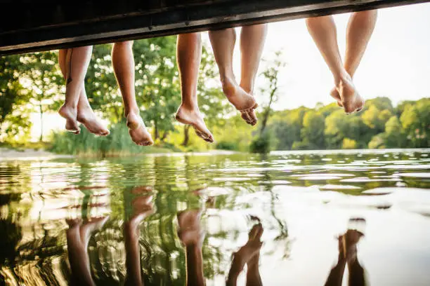 A group of friends legs dangling off a jetty, by the water at a lake together on a sunny afternoon.