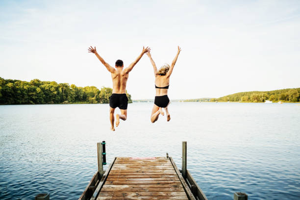 dos amigos saltando juntos de embarcadero en el lago - saltar actividad física fotos fotografías e imágenes de stock