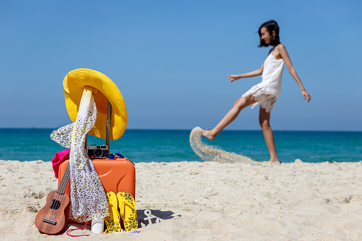 Summer holiday and traveling with orange suitcase and yellow hat, ukulele with woman swimsuit and vintage camera, sandals and sunglasses on the sand beach with happy woman in background