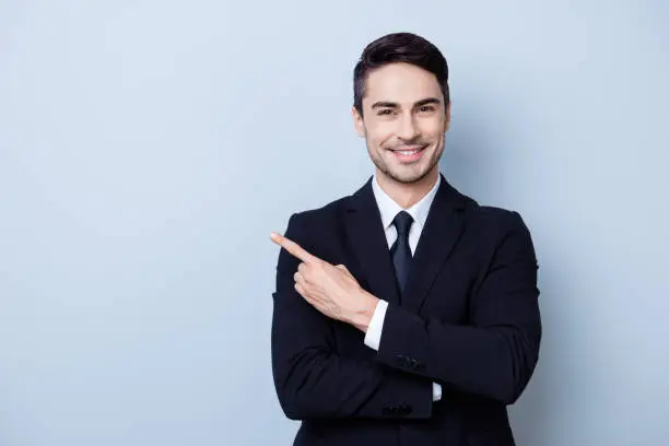 Photo of Close up portrait of young successful brunete  stock-market broker guy on the pure light blue background, he is smiling, wearing suit with tie and is pointing on a copyspace with his finger