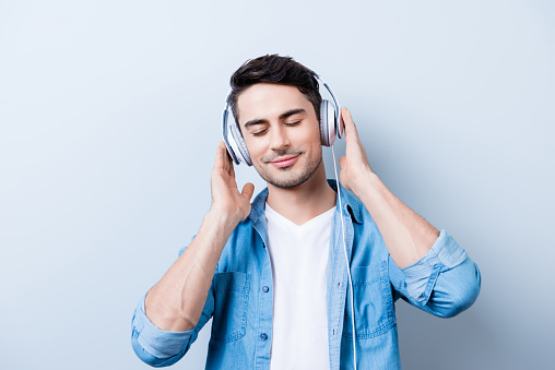 Close up portrait of cheerful young man enjoying listening to his favourite song at his holiday with closed eyes in big white earphones, wearing casual jeans outfit