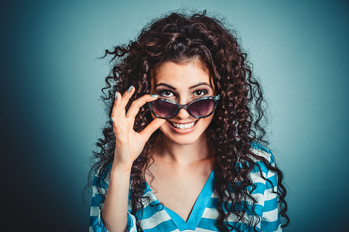 Portrait closeup attractive beautiful happy successful young woman girl holding down sunglasses looking above them smiling at camera isolated blue background wall. Positive human emotion face expression
