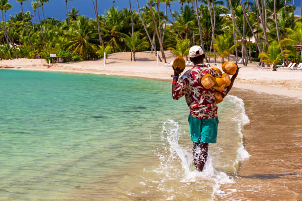 Caribbean coconut saler in walking in a tropical beach of Dominican Republic. SANTO DOMINGO - DOMINICAN REPUBLIC - October 28,2014 : A coconut saler walking by the sea in the famous Juan Dolio Beach with the atmospheric background of palm trees. punta cana stock pictures, royalty-free photos & images
