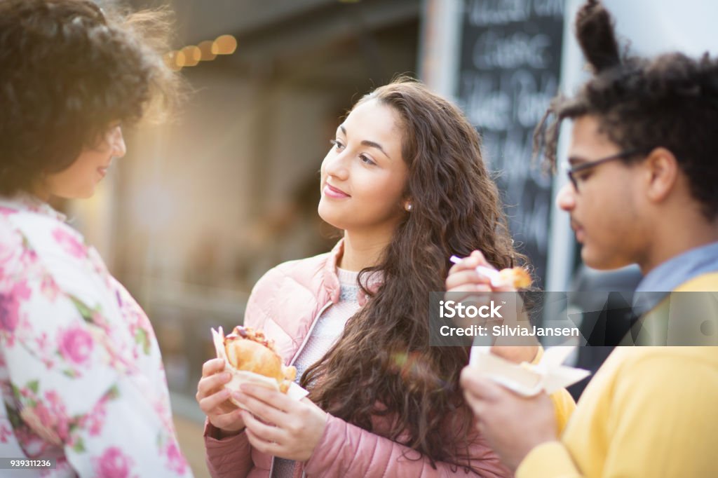 Street Food: friends  eating burger in front of food truck A group of three young adults, two hispanic women and an african man, standing in front of a food truck, eating burger and having fun together 20-29 Years Stock Photo