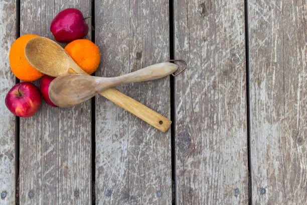 Two serving spoons made of wood and bamboo sit crossed on apples and oranges in a barn wood surface
