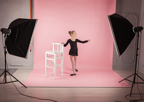 Portrait of young beautiful child girl ballerina standing near chair practicing ballet wearing black tutu dress posing in photo studio pink background, soft box lighting equipment . Copy space image.