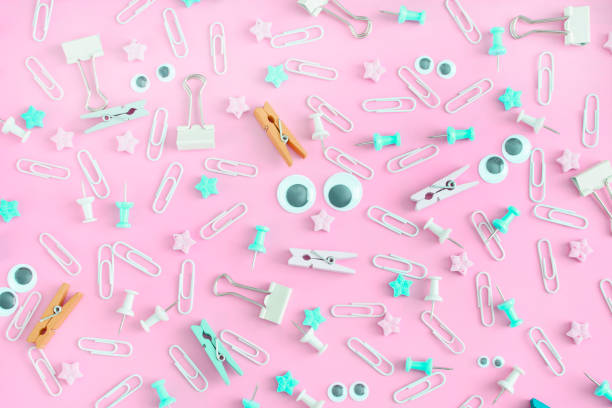 istock A bright picture with scattered stationery. Funny faces with puppet eyes. Clips, clothespins, clerical buttons in a mess on a pink background. 939328532