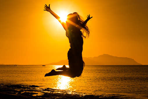 Under the vast orange sky over the Adriatic Sea during sunset in Rovinj,Croatia,a low angle view captures a female tourist standing with arms raised. The scene radiates a sense of joy and wonder,reflecting the magical beauty of the coastal evening