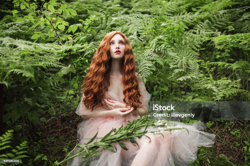 Beautiful pre-raphaelite girl with curly red hair with a flying tulle dress on the background of a green fern Pre-Raphaelite Stock Photo