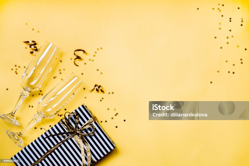 Celebration background - top view of two chrystal champagne glasses, a gift box wrapped in black and white striped paper, ribbons and star shaped golden confetti over yellow background. Copy space. Christmas Stock Photo