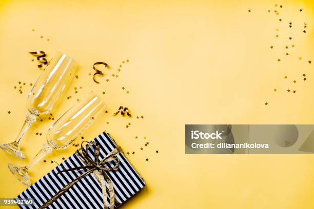 Celebration Background Top View Of Two Chrystal Champagne Glasses A Gift Box Wrapped In Black And White Striped Paper Ribbons And Star Shaped Golden Confetti Over Yellow Background Copy Space Stock Photo - Download Image Now