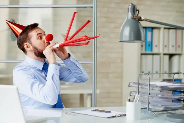 Handsome businessman with beard blowing party horns with the full force in modern office