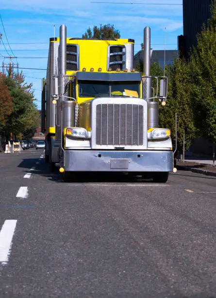 Bright yellow American classic big rig semi truck fleet with freezer semi trailer and tall chrome exhaust pipes running by urban city street for timely delivery of goods to the customers