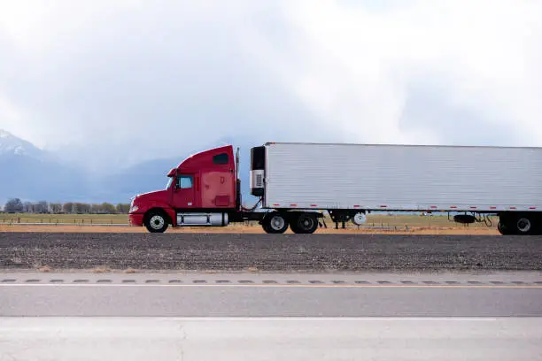 Photo of Red big rig semi truck carry refrigeration semi trailer and going by flat road in Utah