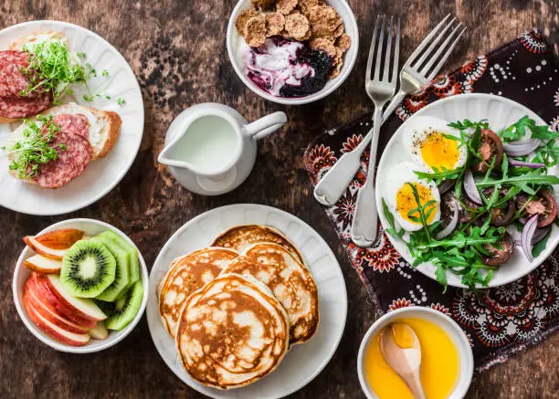 Photo of Greek yogurt with whole grain cereals and berry sauce, pancakes, arugula, cherry tomatoes, boiled eggs salad, kiwi, apples fruit, salami and cream cheese sandwiches on a wooden background, top view. Flat lay breakfast table