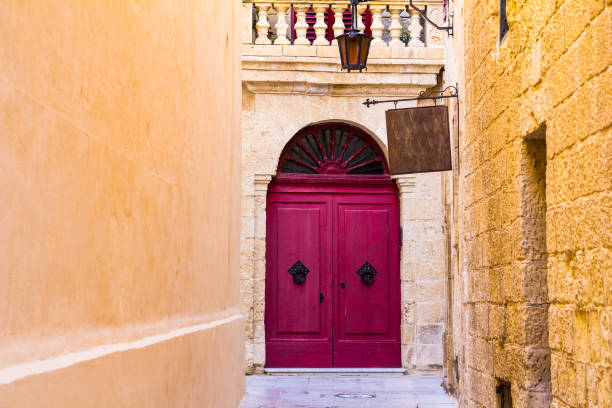 Color image depicting a narrow ancient cobbled street leading to a vibrant purple-colored door in Mdina, an ancient and charming town on the island country of Malta. There is a wooden sign with an arrow pointing in the direction of a tea garden. Lots of room for copy space.