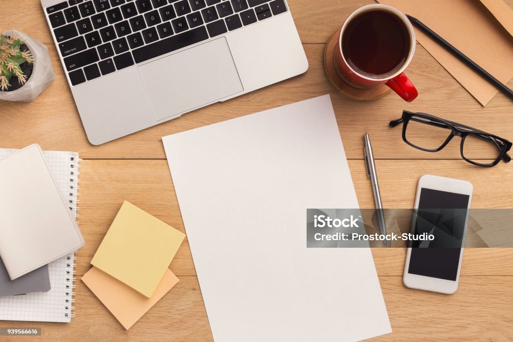 Wooden table with empty paper top view Male workplace with empty sheet of paper for advertisement, smartphone with blank screen, laptop, coffee cup and glasses. Wooden background, top view, mockup, copy space. Desk Stock Photo