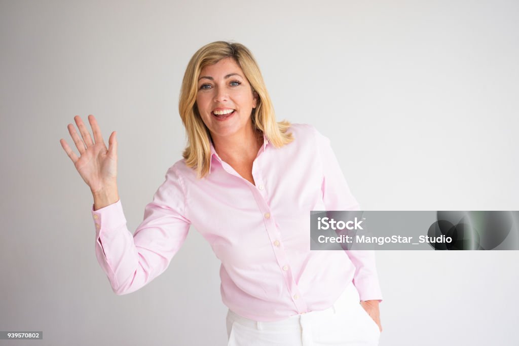 Smiling Pretty Woman Waving with Hand Closeup portrait of smiling middle-aged attractive fair-haired woman looking at camera and waving with her hand. Greeting concept. Isolated front view on grey background. Waving - Gesture Stock Photo