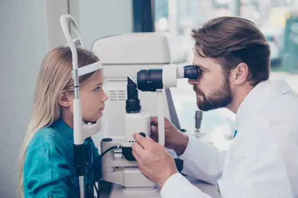 Photo of Health care, medicine, eye sight and technology concept. Focused brunet bearded optometrist with non contact tonometer is checking small blond`s girl patient intraocular pressure at eye clinic