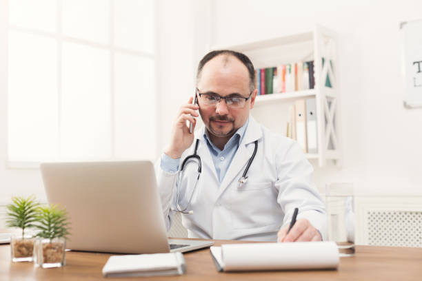 Happy doctor talking on phone with his patient Happy doctor talking on phone with one of his patients and taking notes while working at his office. Always ready to help, online consultation, medicine concept, copy space technology office equipment laboratory stock pictures, royalty-free photos & images