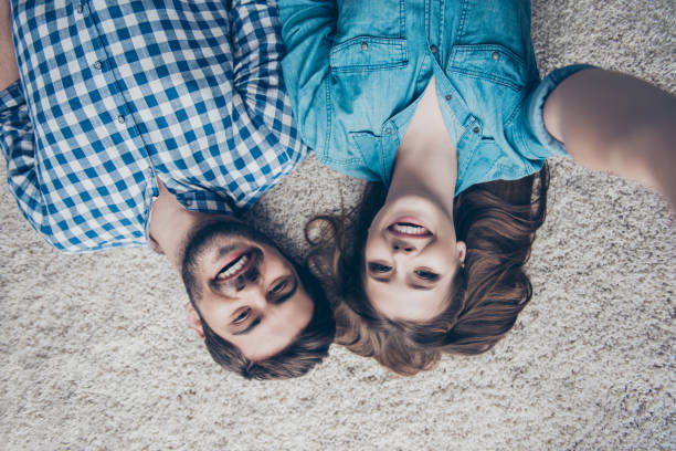 do góry nogami widok zdjęcie dwóch młodych przyjaciół, którzy robią selfie i uśmiechają się. leżą na podłodze na beżowym dywanie w swobodnych strojach w domu - carpet floor lying down people zdjęcia i obrazy z banku zdjęć
