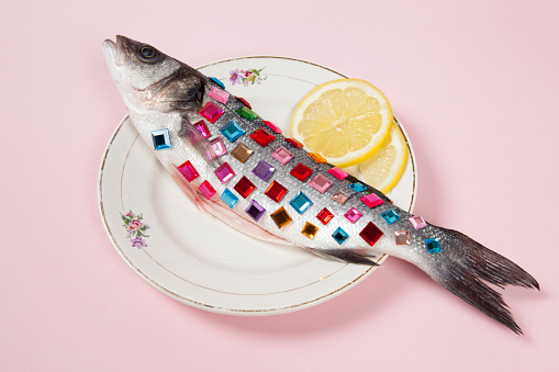 A bass fish covered with colorful precious stones inside a flower plate hidden on a pop pink background. Minimal quirky color still life photography.