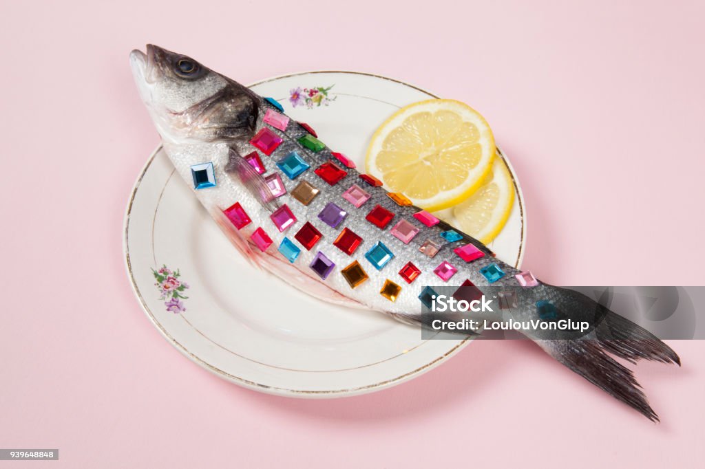 lemon fish pink plate precious gems A bass fish covered with colorful precious stones inside a flower plate hidden on a pop pink background. Minimal quirky color still life photography. Bizarre Stock Photo