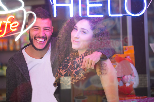 Man shopping in a coffee shop poses with girlfriend at the store window. They are both posing an looking at the camera. They are both Latin in their twenties. He has a modern hair cut with beard. she  has curly hair.