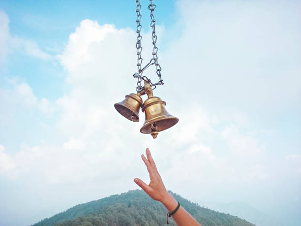 Two temple bells made of brass hanging through chain against cloudy sky and a devotee's hand trying to hit the bell. Place: Kali Ka Tibba, Chail, Himachal Pradesh.
