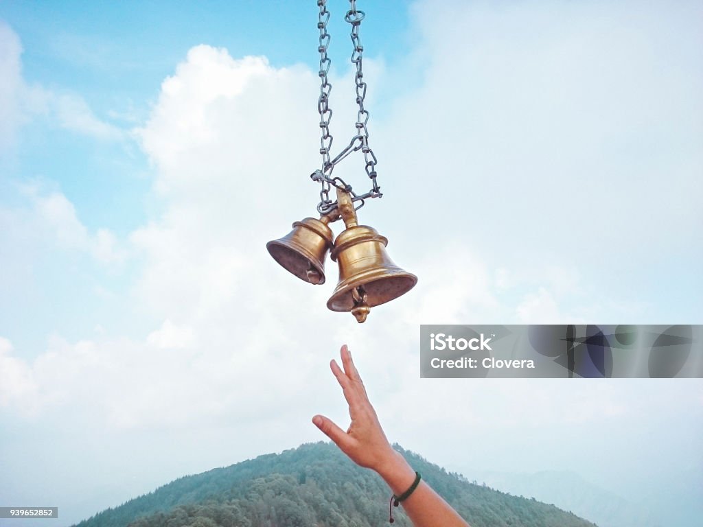 Devotee's hand trying to hit Hindu temple bells. Two temple bells made of brass hanging through chain against cloudy sky and a devotee's hand trying to hit the bell. Place: Kali Ka Tibba, Chail, Himachal Pradesh. Bell Stock Photo