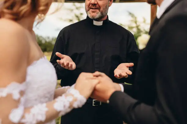 Close up of bride and groom holding each other's hands during wedding ceremony. Priest giving blessings to couple during outdoor wedding ceremony.