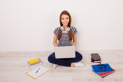 Pretty young female student in striped t-shirt, jeans and headphones sitting on the floor at home with laptop and books and preparing for exams